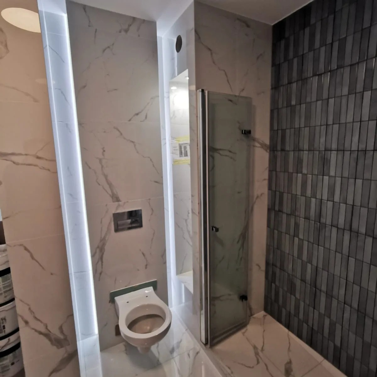 Nowoczesna łazienka z marmurowymi płytkami, podświetleniem LED i szklaną kabiną prysznicową, widok na zawieszony sedes.