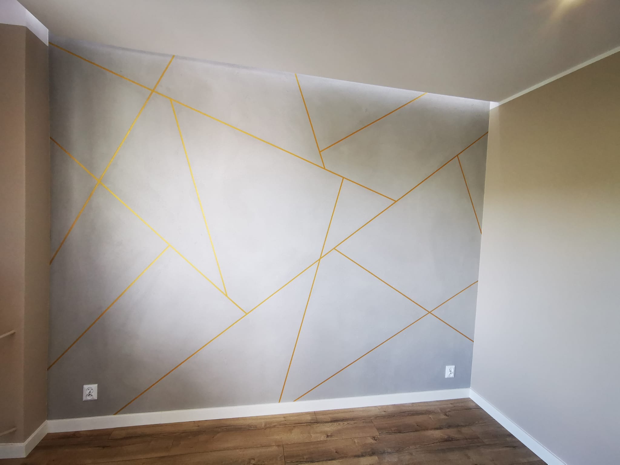 Ściana z gładzi w odcieniach szarości z dekoracyjnymi, złotymi liniami tworzącymi geometryczny wzór, drewnopodobna podłoga, dwie puszki elektryczne na ścianie.
