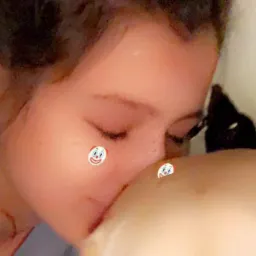 Kobieta z piegami na twarzy delikatnie całuje w głowę niemowlę, oboje mają nałożone filtry z emotikonami klaunów.