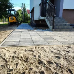 Nowo ułożona, szara kostka brukowa przed domem, obok schodów wejściowych i małej koparki JCB, na piasku widoczne ślady stóp.