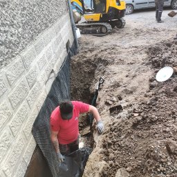 WMO Roboty Ogólnobudowlane - Pracownik w wykopie przy fundamencie budynku, układa czarną folię hydroizolacyjną i membranę kubełkową, w tle minikoparka JCB i zaparkowany samochód dostawczy.