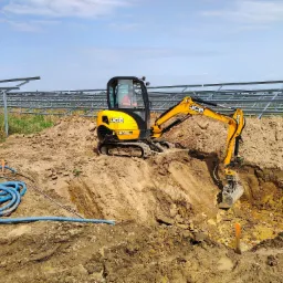 Żółta minikoparka JCB 8026 CTS w trakcie prac ziemnych, wykop pod instalację, w tle konstrukcja farmy fotowoltaicznej i błękitne niebo.