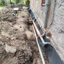 WMO Roboty Ogólnobudowlane - Wykop fundamentowy z widoczną izolacją pionową z folii kubełkowej, rurami drenażowymi i instalacją odprowadzającą wodę, zarys budynku w tle i koparka w oddali.