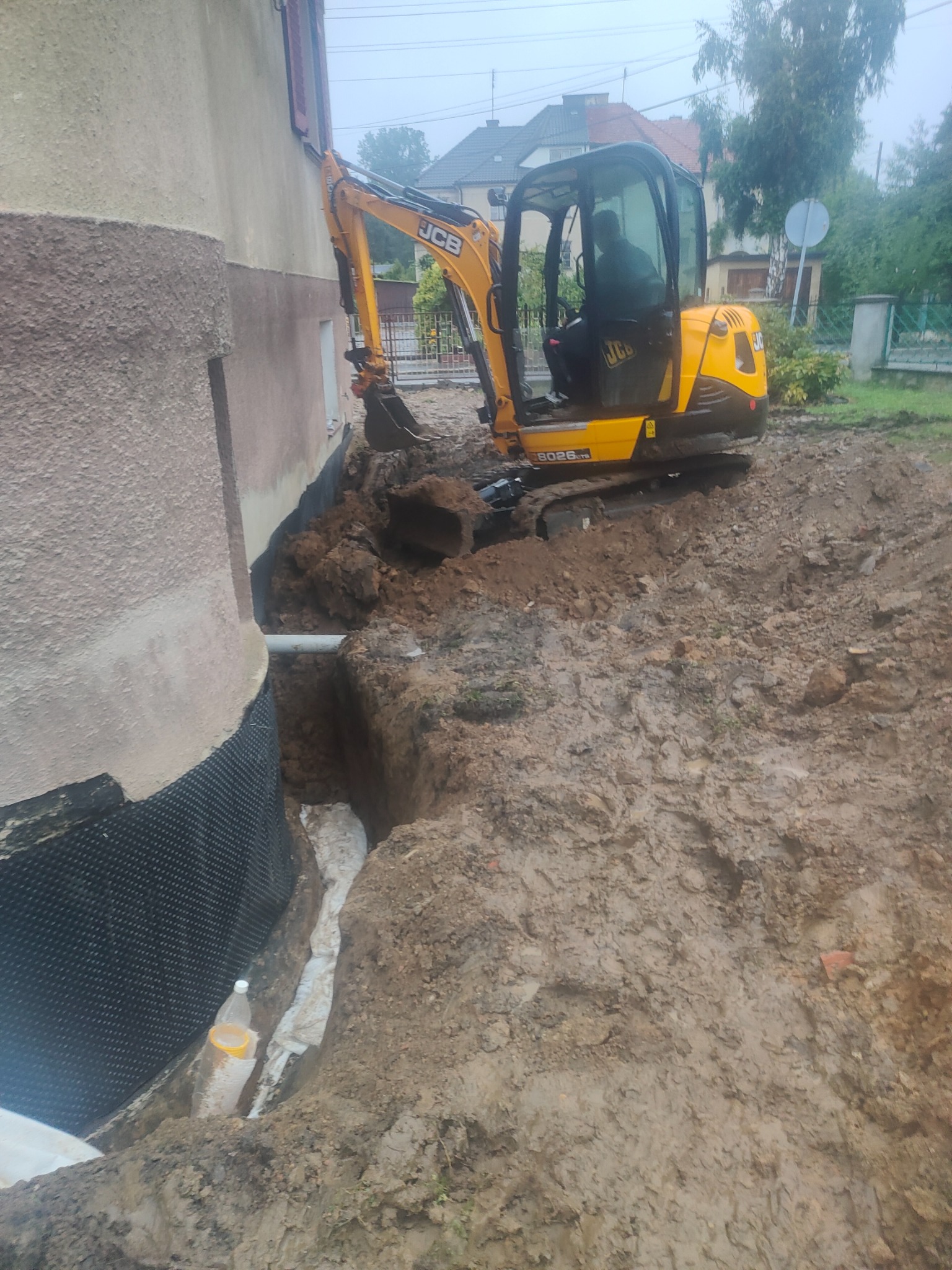 Żółta mini koparka JCB obok fundamentu budynku z wykonaną izolacją pionową, wykopany rów, w którym ułożona jest rura drenażowa owinięta geowłókniną.
