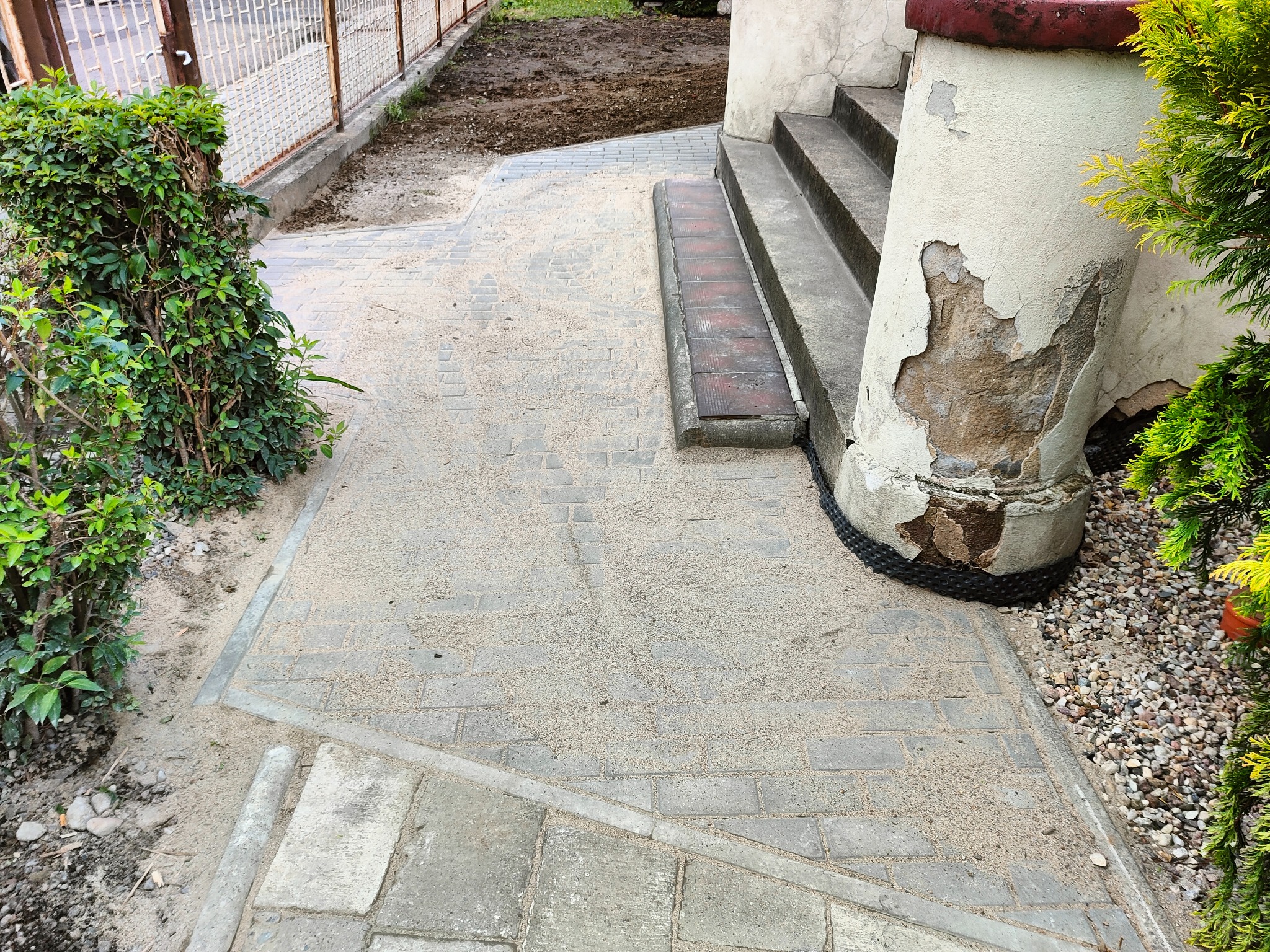 Ułożona szara kostka brukowa z piaskiem na podwórku, obok schodów z uszkodzonym filarem i roślinnością.