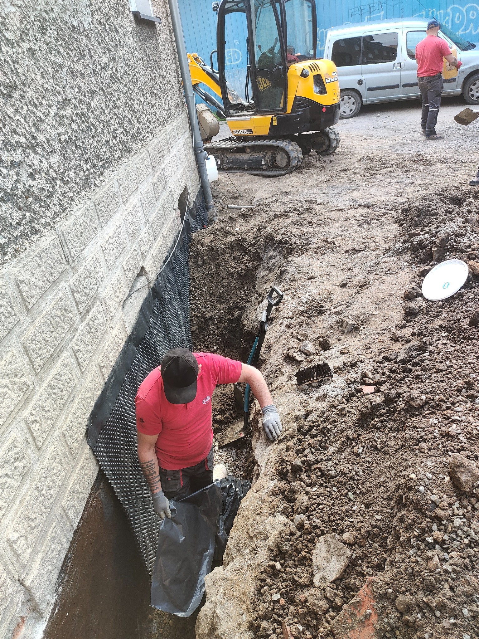 Pracownik w wykopie przy fundamencie budynku, układa czarną folię hydroizolacyjną i membranę kubełkową, w tle minikoparka JCB i zaparkowany samochód dostawczy.