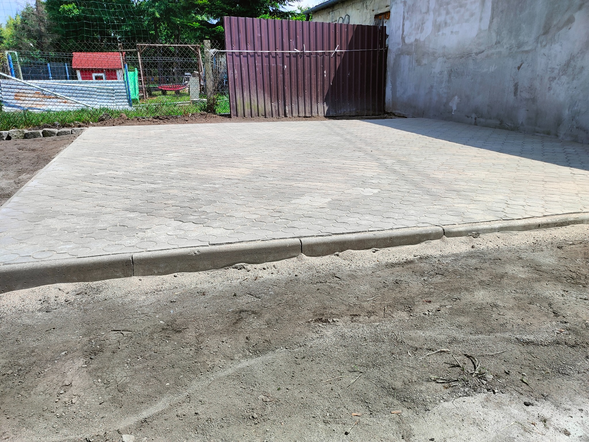 Ułożona szara kostka brukowa na posesji, obrzeża betonowe, w tle siatka ogrodzeniowa i blaszana brama.