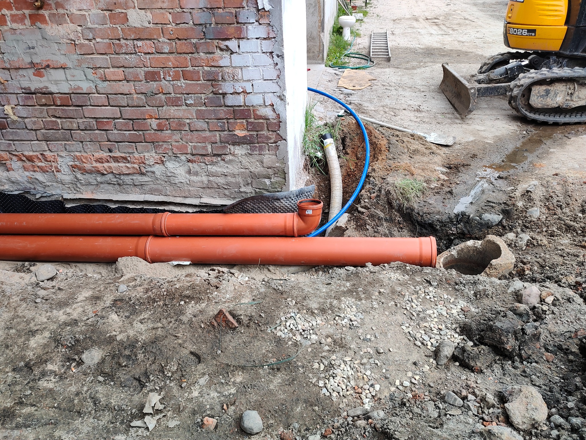 Pomarańczowe rury kanalizacyjne ułożone na gruncie obok fundamentu budynku z cegły, w tle wykop i mini koparka.
