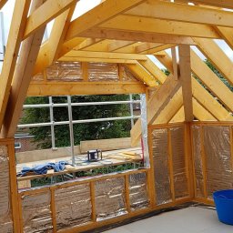 Hubert Szymczak - Wnętrze poddasza w trakcie adaptacji: drewniana konstrukcja dachu, ściany z izolacją pokrytą folią, podłoga z płyt, rusztowanie widoczne przez okno.