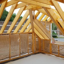 Hubert Szymczak - Surowe drewniane krokwie i izolacja folią aluminiową na ścianach poddasza w trakcie adaptacji, widok z wnętrza, drabina na pierwszym planie.