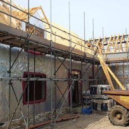 Hubert Szymczak - Budowa domu jednorodzinnego w trakcie realizacji, widoczne rusztowania, ocieplenie ścian folią aluminiową i drewniana konstrukcja dachu, z boku stoi żółta taczka z kołami.