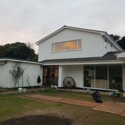 Hubert Szymczak - Biały dom z fakturą na elewacji w trakcie prac wykończeniowych, widoczne puszki z farbą, narzędzia i drewniane płyty na trawniku, zachodzące słońce odbija się w oknach.