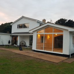 Hubert Szymczak - Dobudowa do domu z dużymi oknami, dwóch mężczyzn przenosi płytę, widoczne elementy wykończeniowe na zewnątrz i wewnątrz.