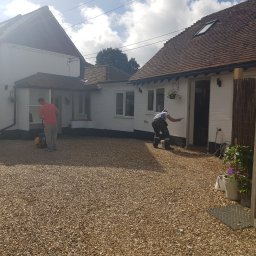 Hubert Szymczak - Dwaj mężczyźni pracują na zewnątrz przy białym, ceglanym domu z brązowym dachem; jeden maluje ścianę, drugi używa zagęszczarki w pobliżu żwirowego podłoża.