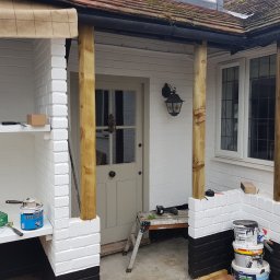 Hubert Szymczak - Biała, ceglana weranda w trakcie malowania z drewnianymi słupami, widoczne narzędzia malarskie i puszki z farbą, czarny pas u dołu ściany.