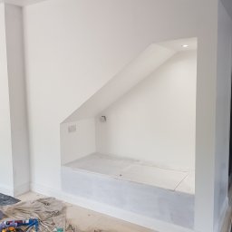 Hubert Szymczak - Wnęka w ścianie po malowaniu na biało, z widocznym oświetleniem punktowym, gładkimi ścianami i narzędziami malarskimi rozłożonymi na podłodze.