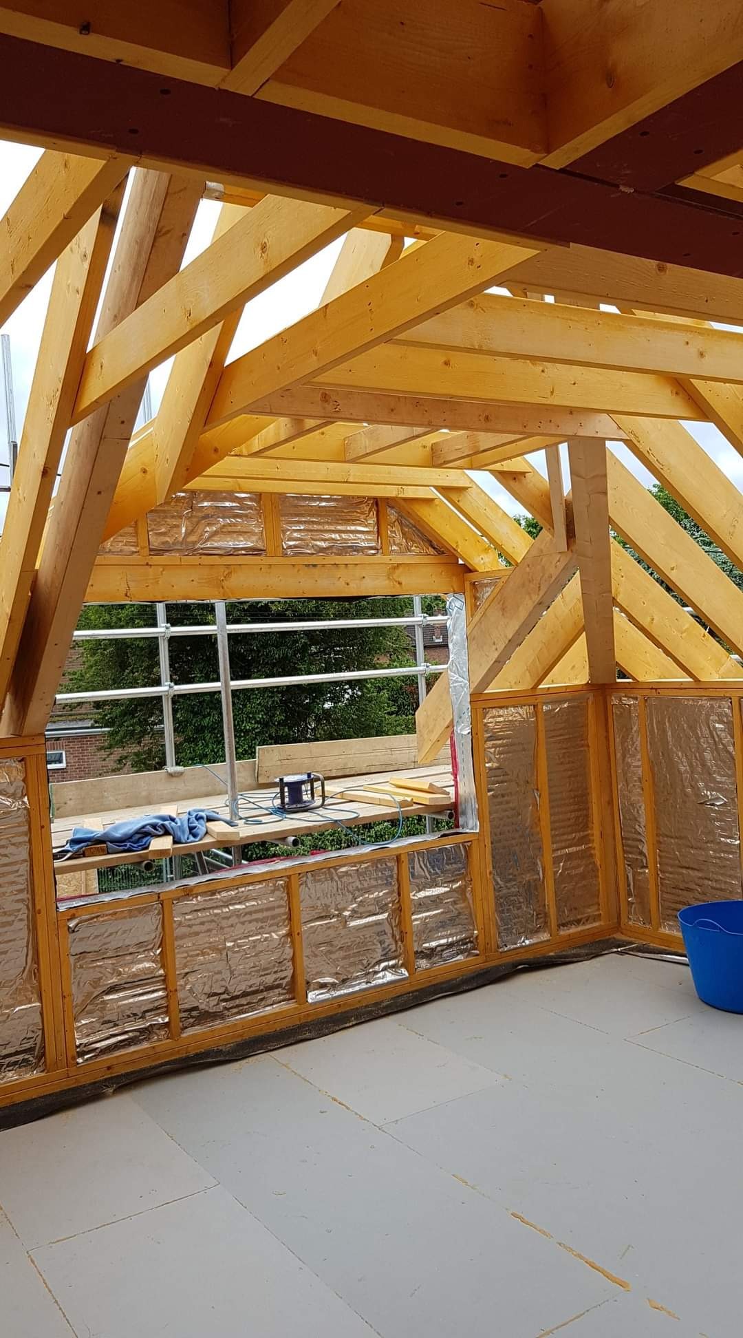 Wnętrze poddasza w trakcie adaptacji: drewniana konstrukcja dachu, ściany z izolacją pokrytą folią, podłoga z płyt, rusztowanie widoczne przez okno.