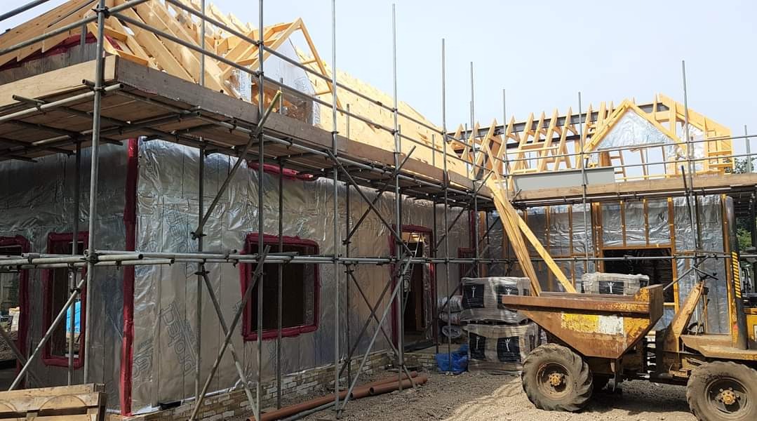 Budowa domu jednorodzinnego w trakcie realizacji, widoczne rusztowania, ocieplenie ścian folią aluminiową i drewniana konstrukcja dachu, z boku stoi żółta taczka z kołami.