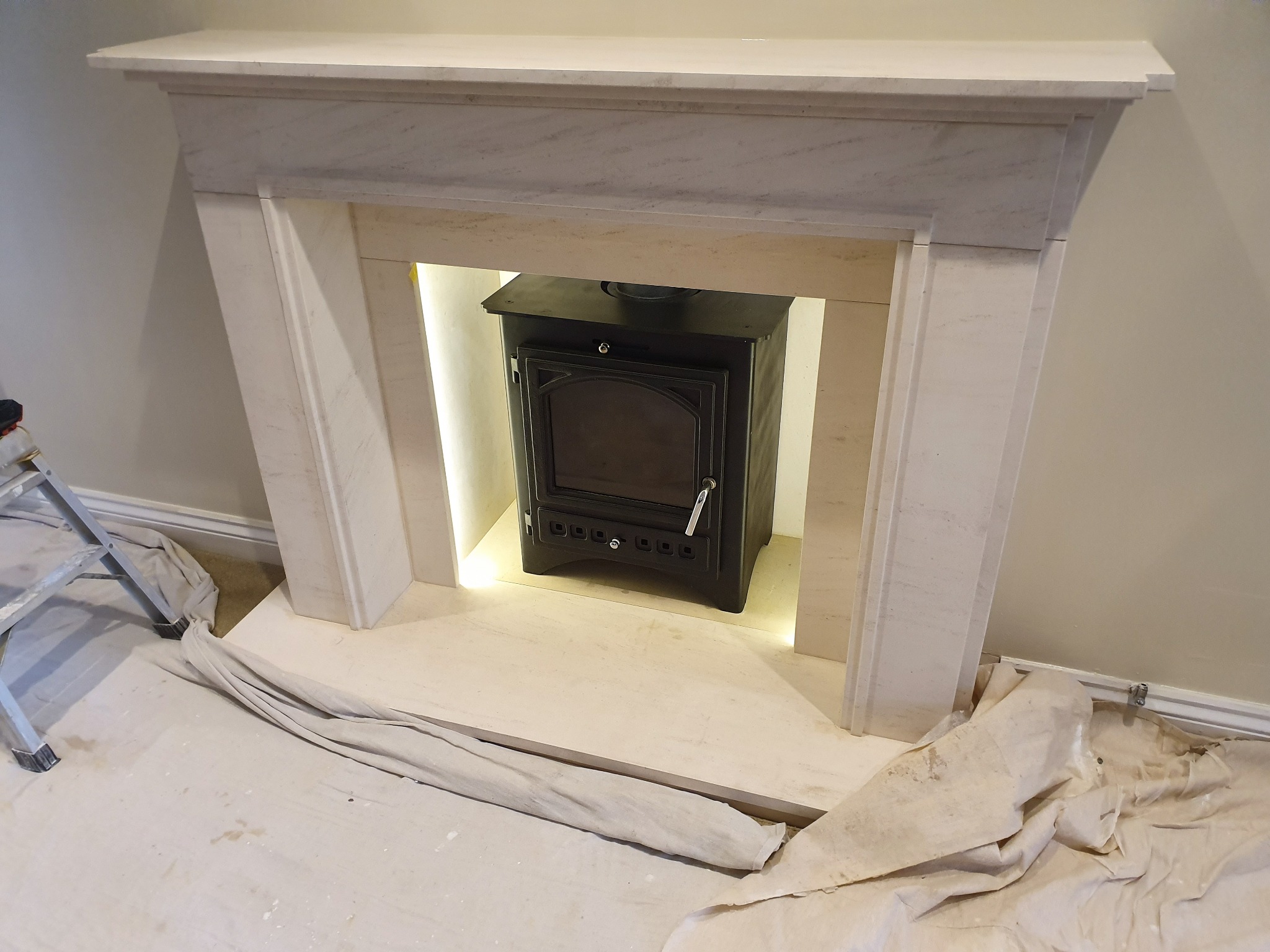 Marmurowy kominek z czarnym piecem na drewno, oświetlenie LED w niszy, podłoga zabezpieczona materiałem malarskim, drabina malarska w tle.