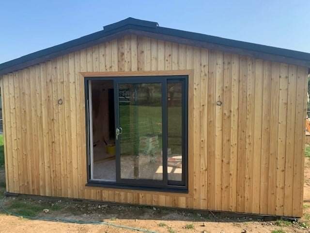 Nowoczesny, drewniany domek letniskowy z przesuwnymi drzwiami, wykończony pionowymi deskami w naturalnym odcieniu drewna, widoczny z zewnątrz na tle zielonego trawnika.