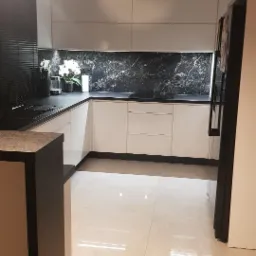 Nowoczesna kuchnia z białymi frontami, czarnym blatem i marmurowym backsplash'em. Minimalistyczne oświetlenie sufitowe i jasna podłoga. Widok na fragment lodówki.