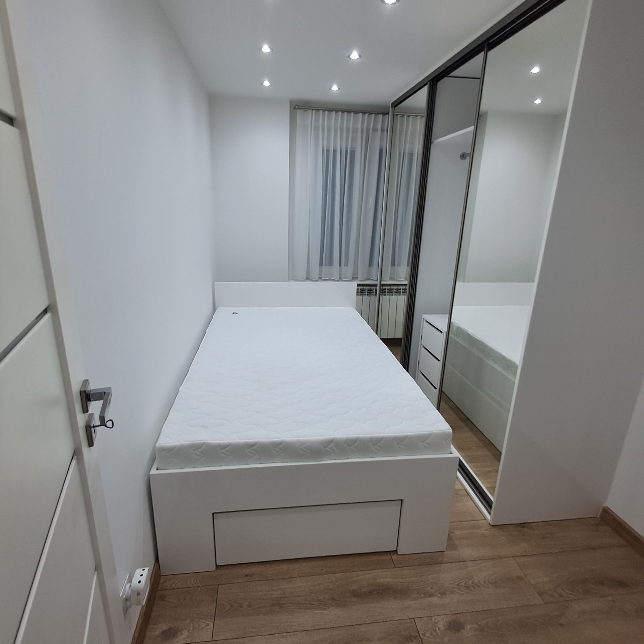 Biały pokój z łóżkiem z szufladą i szafą przesuwną z lustrami. Minimalistyczny design, drewniana podłoga, jasne ściany i okno z zasłoną.