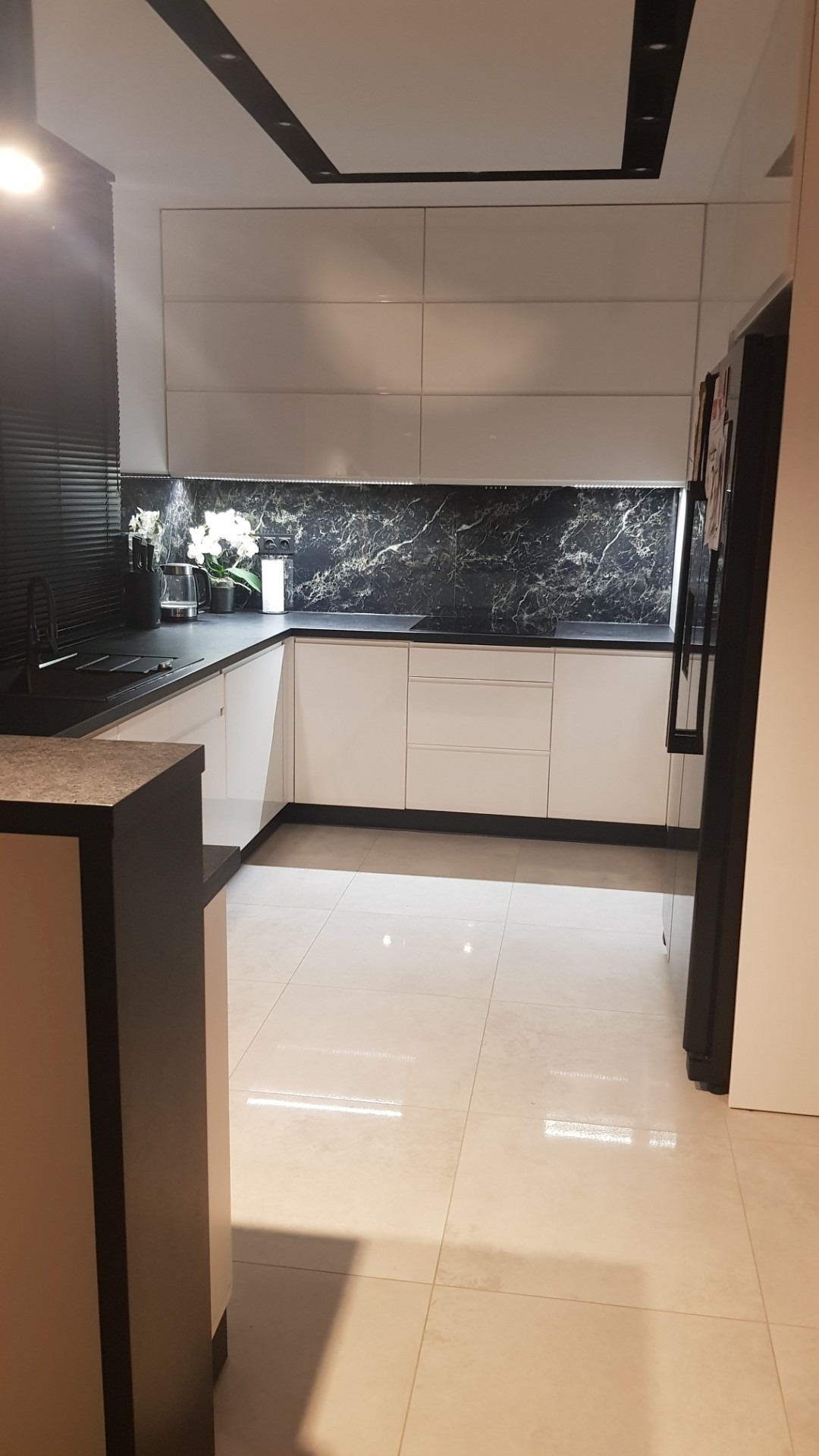 Nowoczesna kuchnia z białymi frontami, czarnym blatem i marmurowym backsplash'em. Minimalistyczne oświetlenie sufitowe i jasna podłoga. Widok na fragment lodówki.