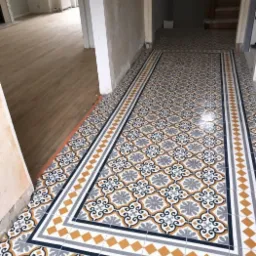 Wzorzysta, nowo ułożona podłoga z płytek ceramicznych w przedpokoju; wzór w kolorach szarym, białym, granatowym i pomarańczowym, z dekoracyjnym obramowaniem; w tle widoczne wejścia do pomieszczeń...