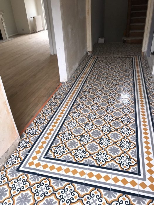 Wzorzysta, nowo ułożona podłoga z płytek ceramicznych w przedpokoju; wzór w kolorach szarym, białym, granatowym i pomarańczowym, z dekoracyjnym obramowaniem; w tle widoczne wejścia do pomieszczeń...