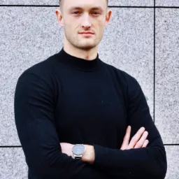 Młody mężczyzna w czarnym golfie, z założonymi rękami i zegarkiem na tle szarej, teksturowanej ściany.