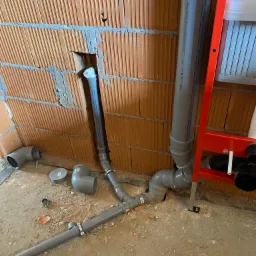 Szare rury kanalizacyjne z kształtkami, częściowo zamontowane w surowej ścianie z cegły dziurawki, widoczny stelaż podtynkowy WC w kolorze czerwonym.