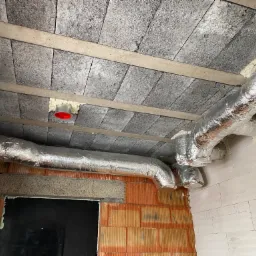 Wnętrze w trakcie budowy z widocznymi, izolowanymi rurami wentylacyjnymi pod sufitem z betonowych bloczków, zamocowanymi pianką montażową. Na ścianach widoczne bloczki z betonu komórkowego i cegły.