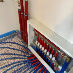 Rozdzielacz ogrzewania podłogowego z niebieskimi rurkami ułożonymi na folii refleksyjnej, widoczne pionowe rury w czerwonej izolacji, świeżo położone instalacje w surowym stanie.