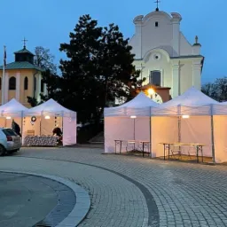Białe namioty ustawione na brukowanym placu przed budynkiem kościoła w szarówce, widoczne oświetlenie wewnątrz namiotów i osoby przygotowujące stoiska.