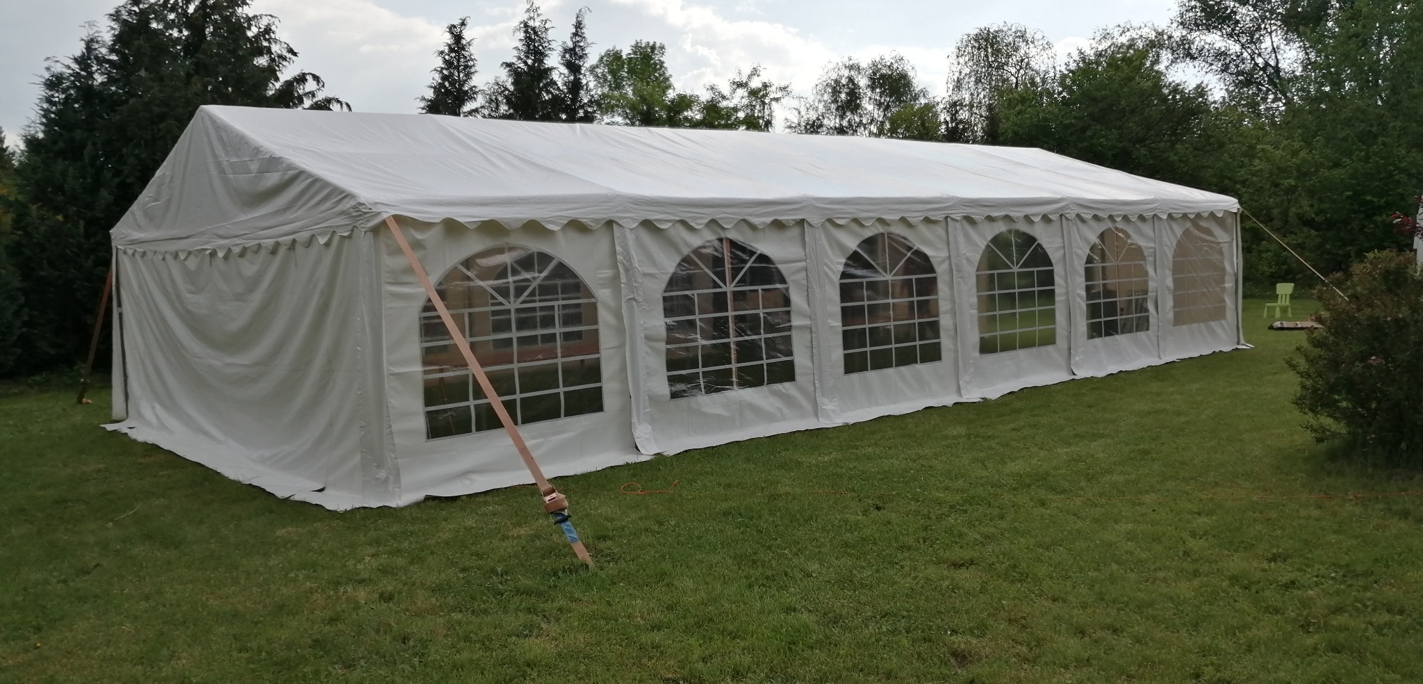 Rozłożony biały namiot imprezowy z oknami na trawniku, zabezpieczony pasami mocującymi, z zielonym krzesłem ogrodowym widocznym w tle.