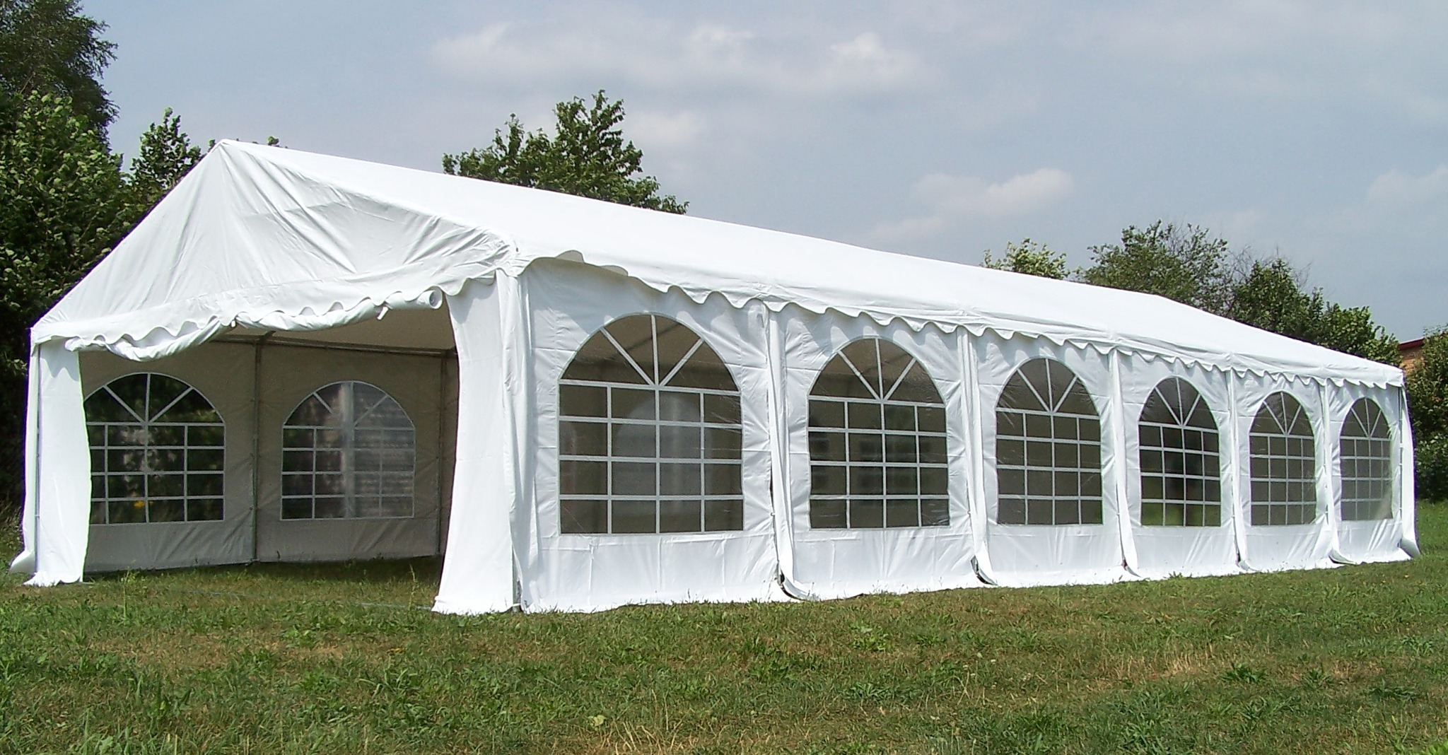 Duży, biały namiot imprezowy z oknami w kształcie łuków, ustawiony na trawie, widoczne drzewa w tle.
