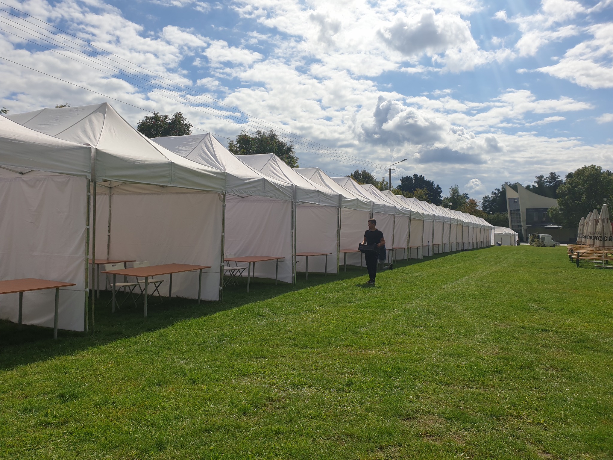 Rząd białych namiotów z rozstawionymi stołami na zielonej trawie, błękitne niebo z chmurami w tle, osoba przechodząca obok namiotów.