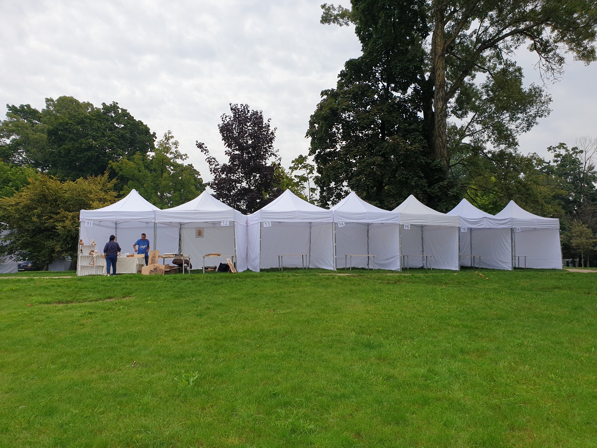 Rząd białych namiotów z numerami na trawiastym terenie, widoczne osoby w jednym z namiotów, drzewa w tle.