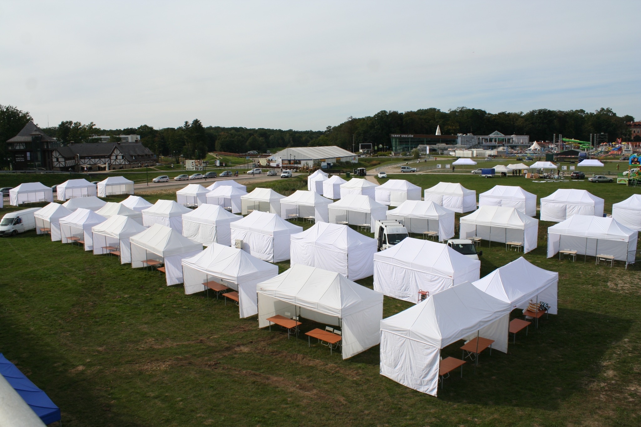 Widok z góry na rozstawione białe namioty z drewnianymi stołami i krzesłami na zielonej trawie, w tle budynki i drzewa.