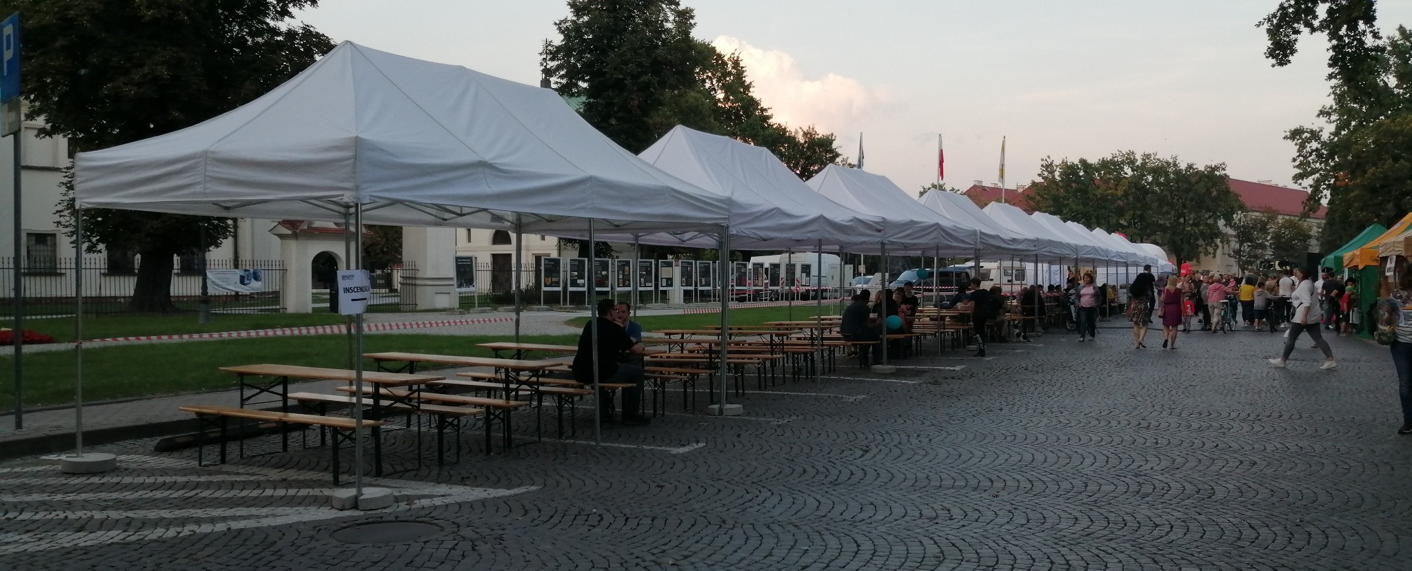 Rząd białych namiotów z drewnianymi stołami i ławkami ustawiony na brukowanej ulicy podczas plenerowej imprezy, w tle budynek i spacerujący ludzie.