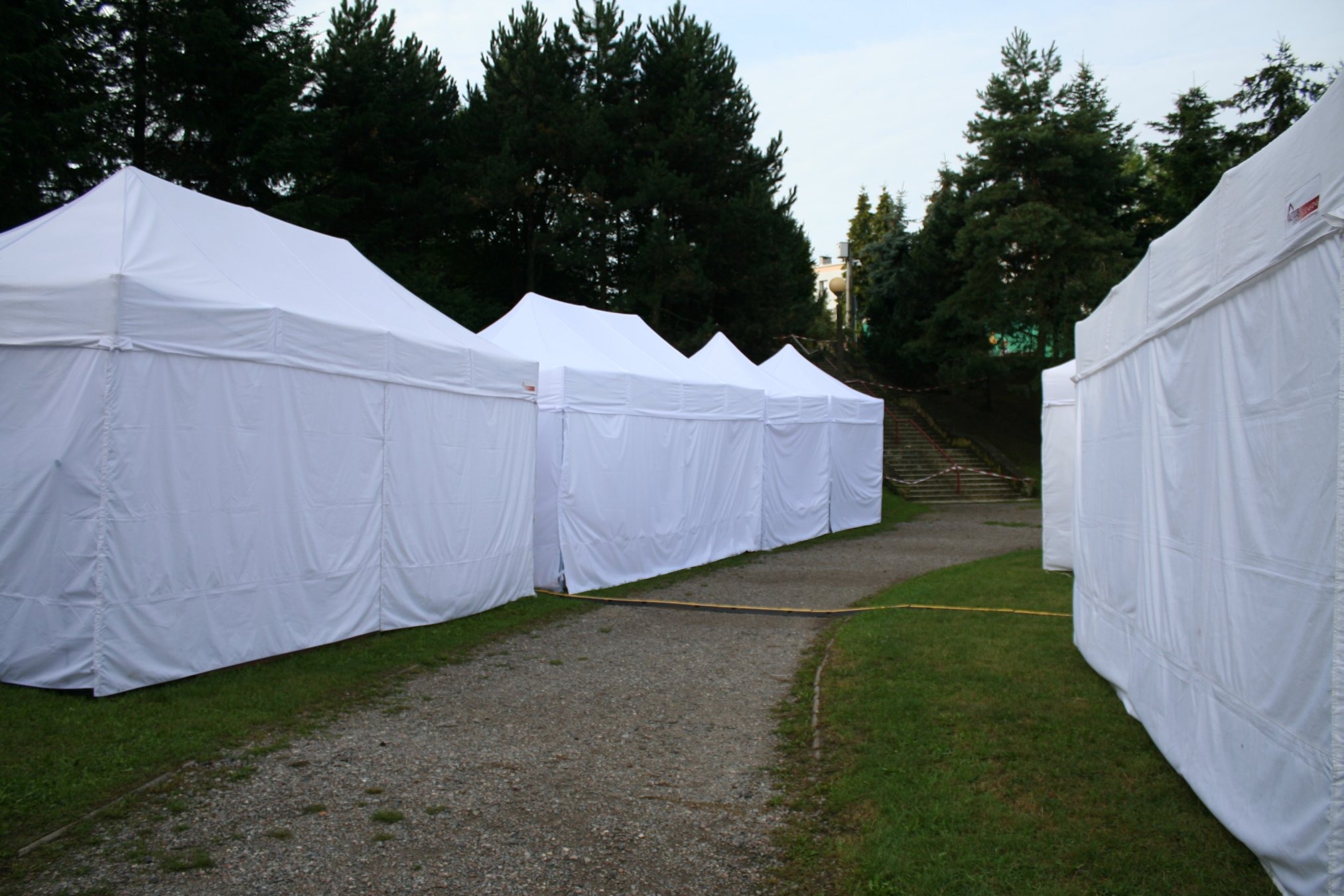 Rząd białych namiotów ustawionych wzdłuż ścieżki na trawiastym terenie, w tle widoczne drzewa i schody.