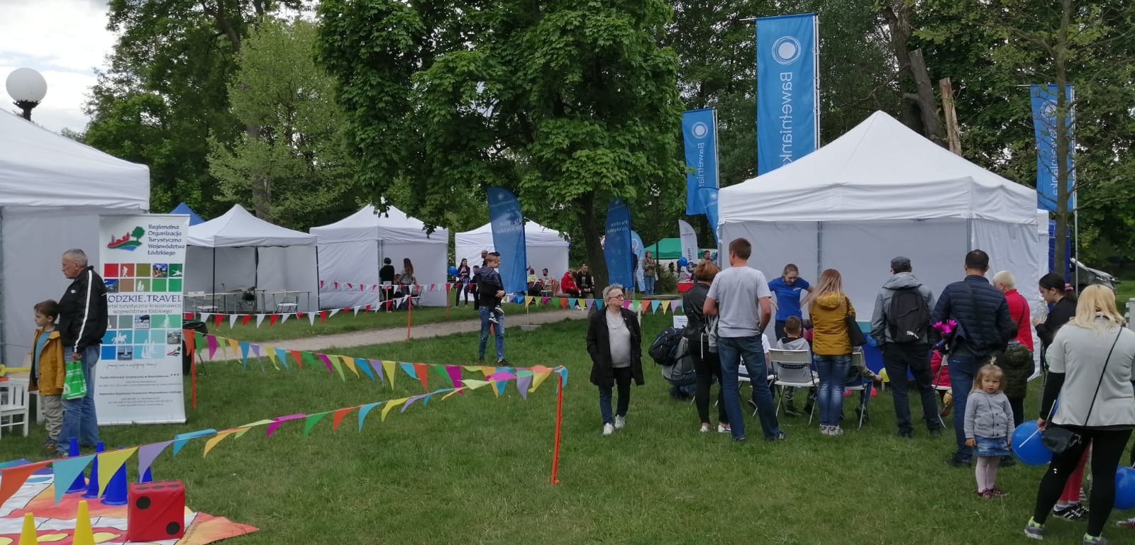 Targi plenerowe z białymi namiotami, flagami reklamowymi i kolorowymi girlandami, widoczni ludzie w różnym wieku.