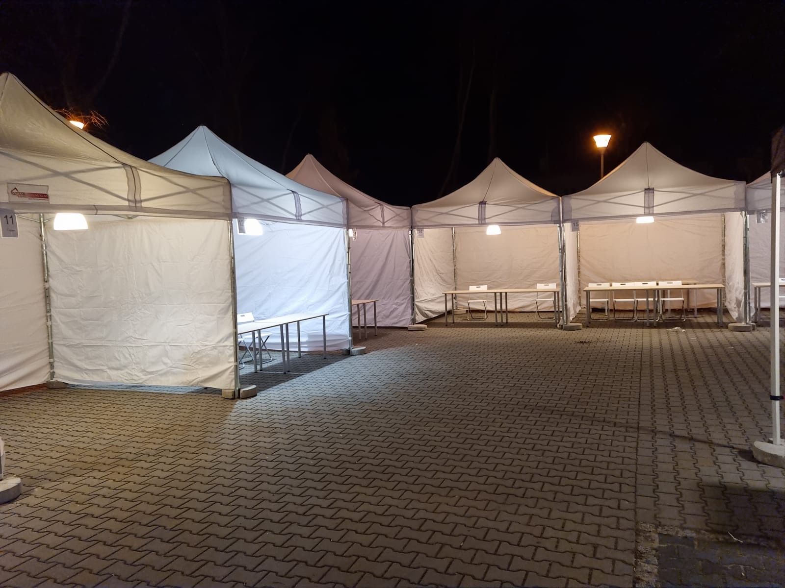Białe namioty ustawione w rzędzie na zewnątrz, oświetlone lampami wewnątrz, z widocznymi stołami i krzesłami, sfotografowane nocą na brukowanym placu.