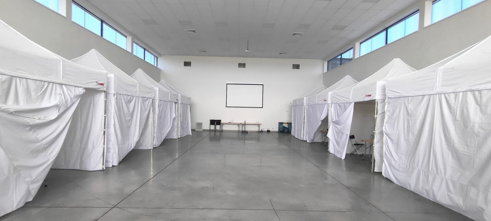 Wnętrze hali z rzędem białych namiotów ustawionych po obu stronach, widoczny ekran projekcyjny i stoliki z krzesłami w tle.
