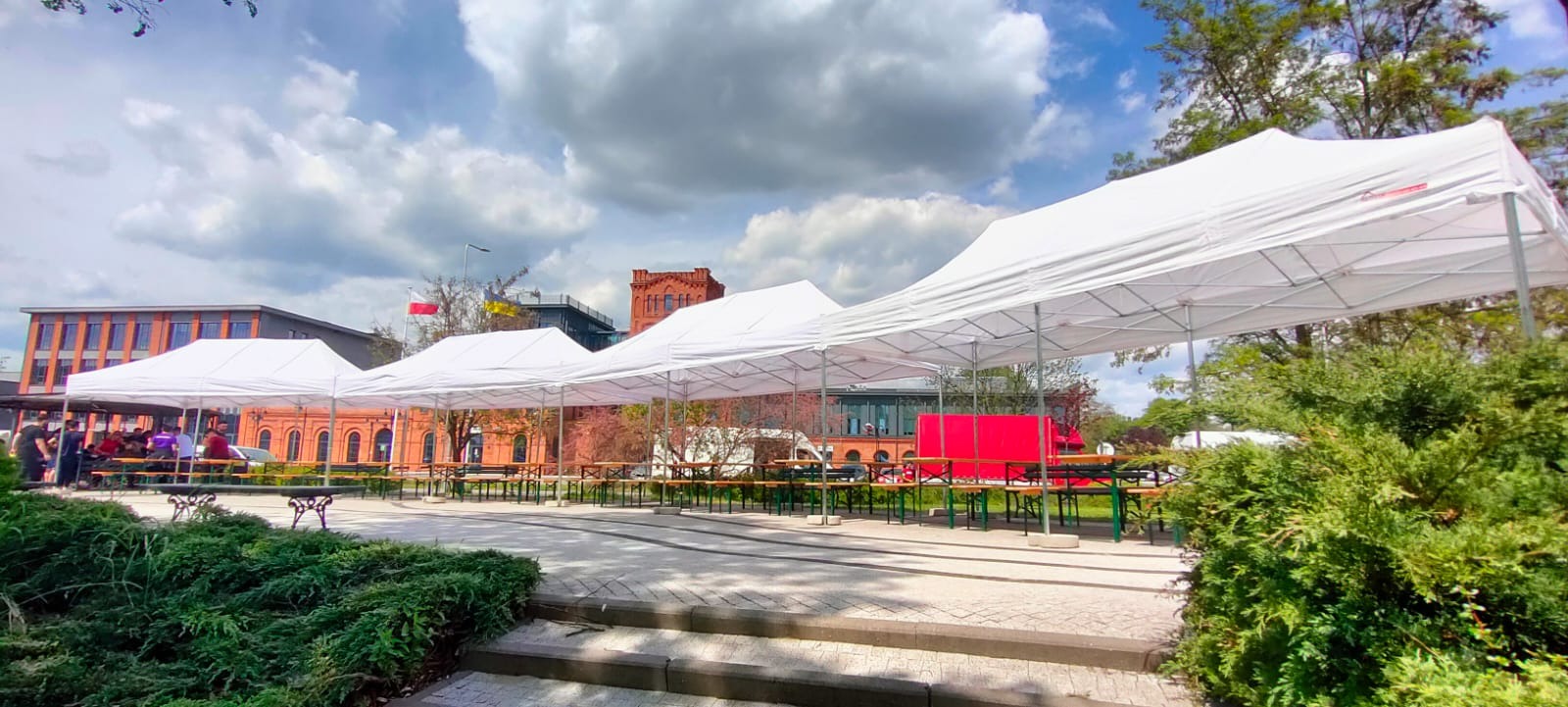 Rozstawione białe namioty z wyposażeniem (stoły i ławki) na zewnątrz, w tle budynki z czerwonej cegły i pochmurne niebo.