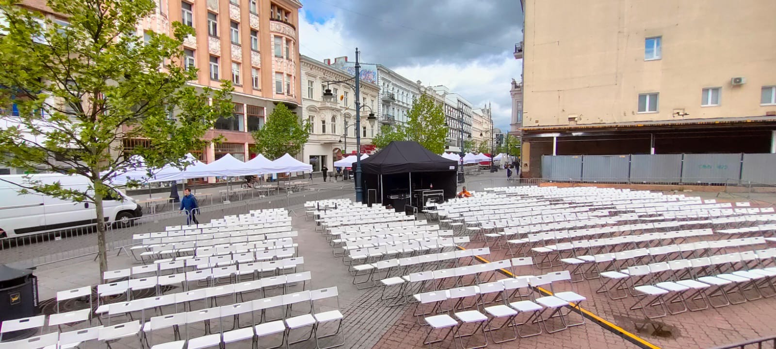 Przygotowania do wydarzenia plenerowego w mieście: rzędy białych krzeseł, czarny namiot z nagłośnieniem, białe namioty w tle, budynki miejskie, pochmurne niebo.