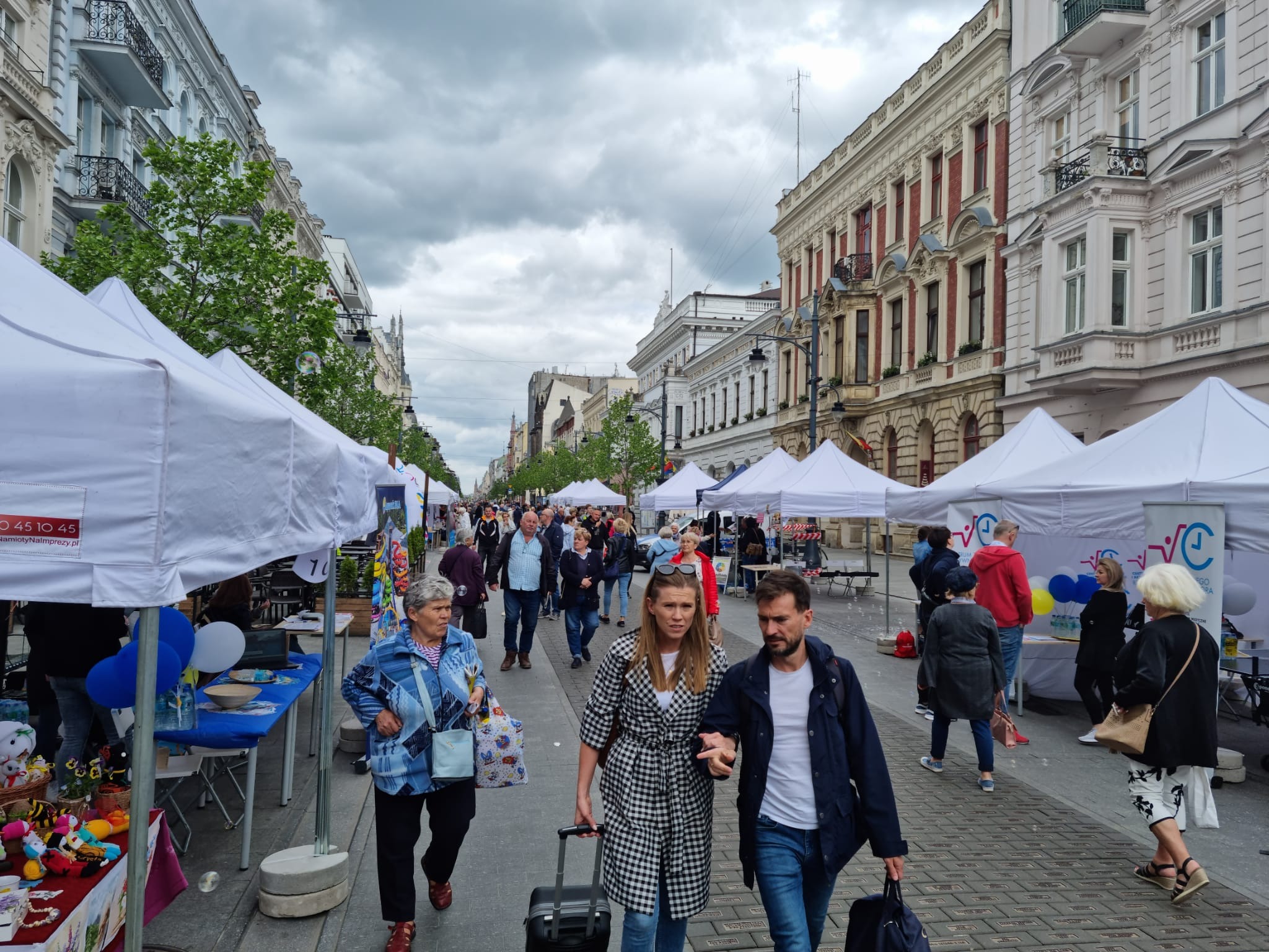 Ulica z białymi namiotami wystawienniczymi po obu stronach, przechodnie spacerują po brukowanej drodze, budynki w tle