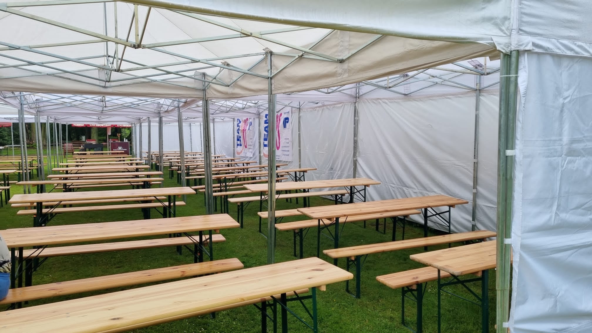 Rozstawione rzędy drewnianych stołów i ławek pod białym namiotem imprezowym na trawie, przygotowane na wydarzenie.