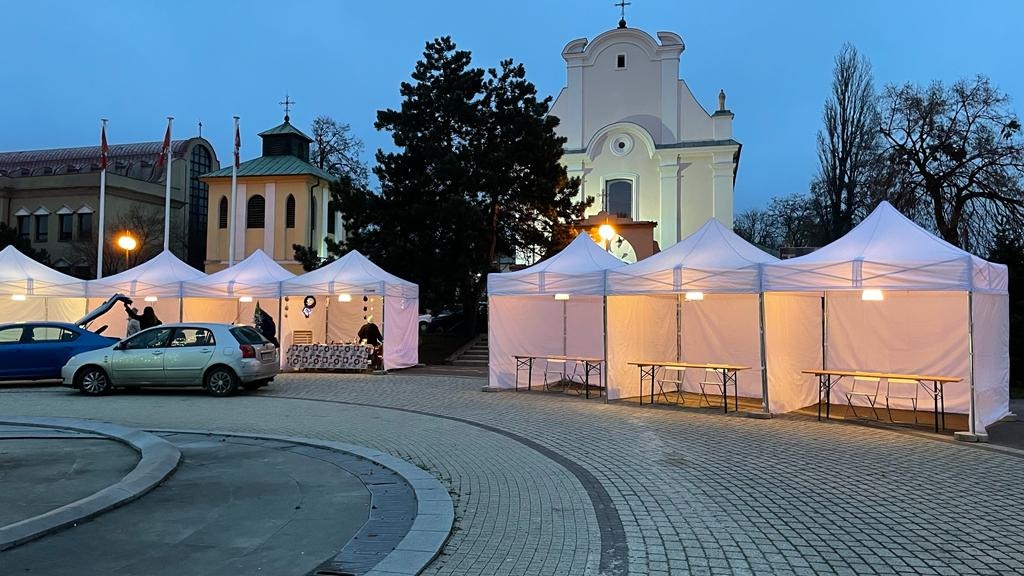 Białe namioty ustawione na brukowanym placu przed budynkiem kościoła w szarówce, widoczne oświetlenie wewnątrz namiotów i osoby przygotowujące stoiska.