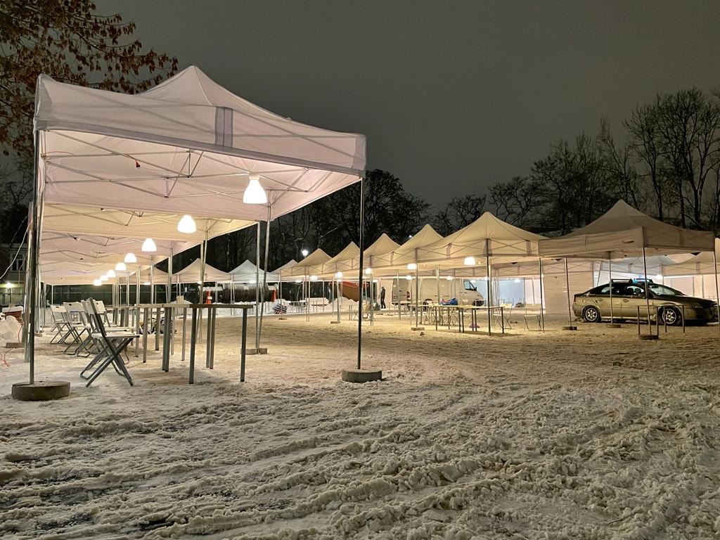 Białe namioty ustawione w rzędzie na śniegu, oświetlone lampami, z samochodami w tle, sceneria nocna.